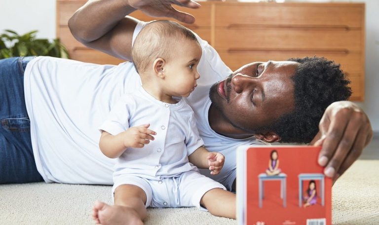 Baby sitting up and being supported by a man looking at a book by Lovevery