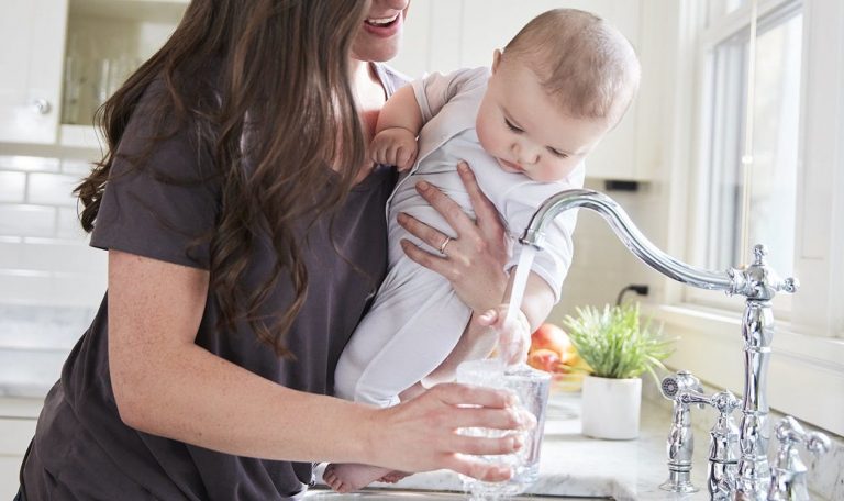 Woman holding baby while pouring water into a glass from the kitchen sink