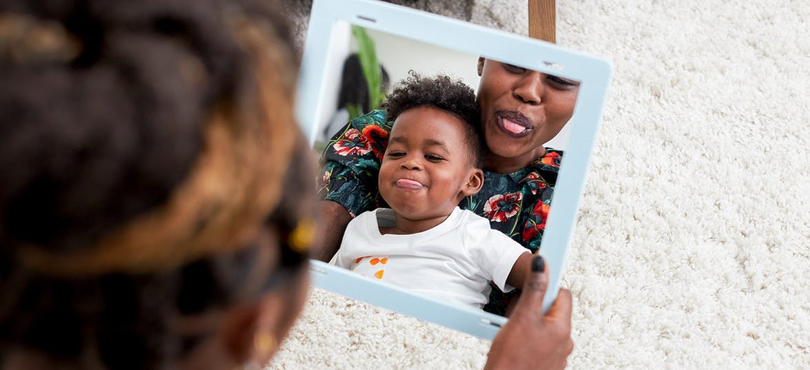 Woman with a toddler in her lap looking into a mirror sticking out their tongues