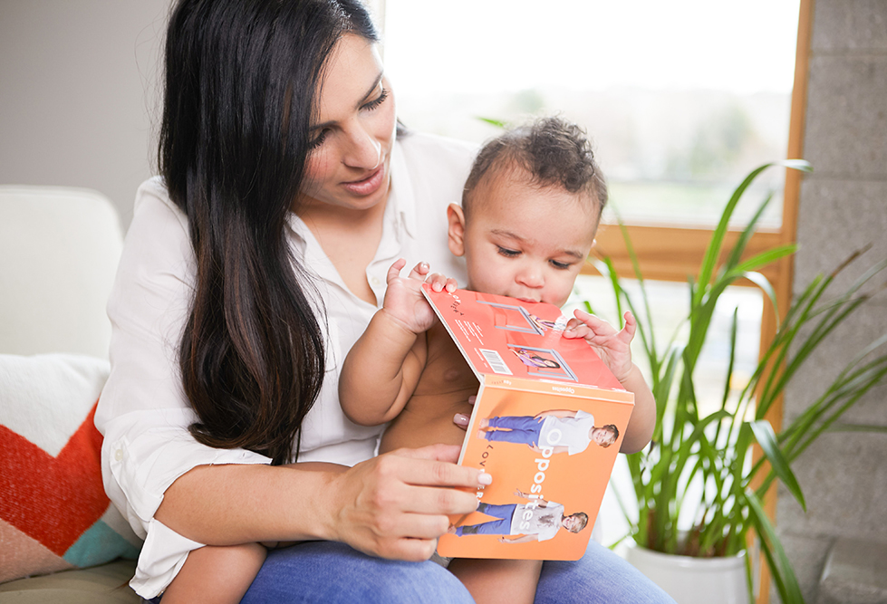 Toddler sitting on a woman's lap while holding the book Opposites by Lovevery