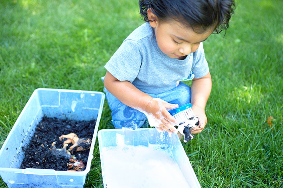 Toddler playing outside with a container of dirt with animal figurines and they're cleaning them off