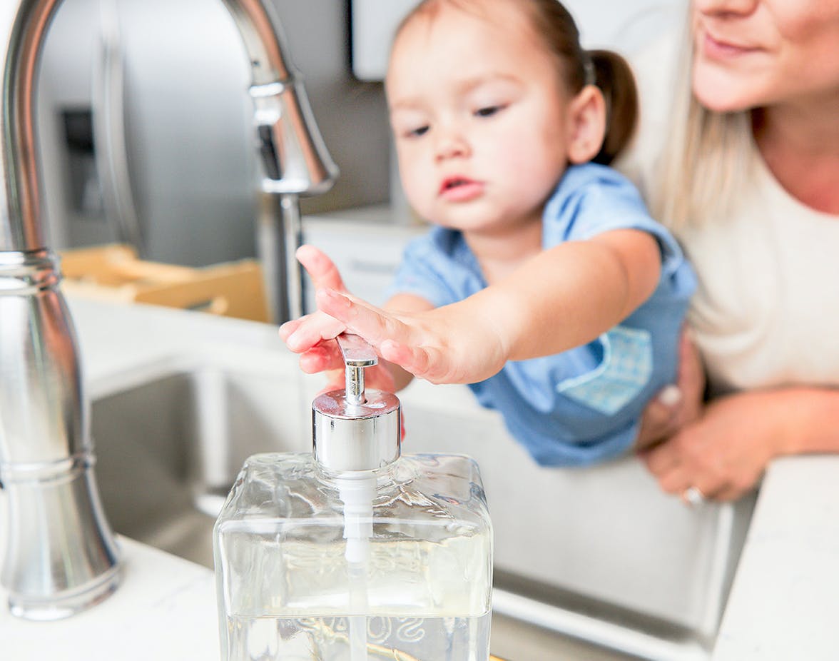 Young child getting soap from a dispenser next to a kitchen sink. 