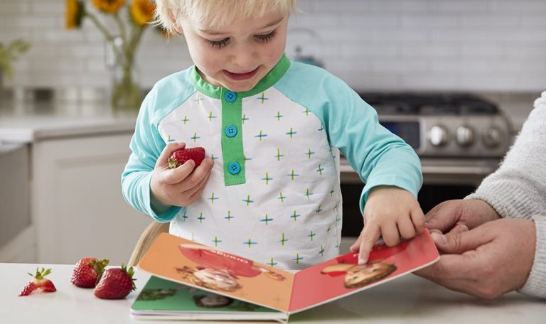 Toddler holding a strawberry and looking at a book by Lovevery