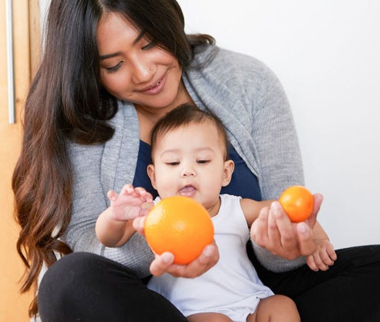 Baby sitting on a woman's lap looking at two different size oranges that she's holding