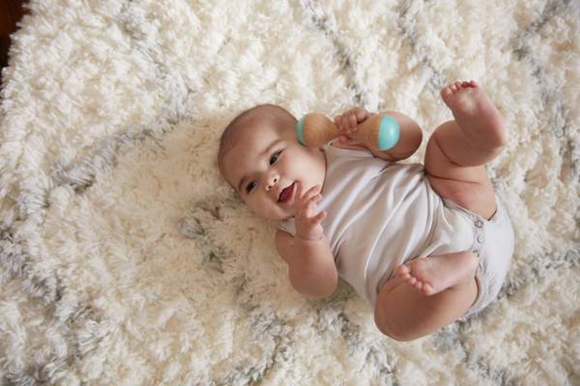 Baby laying on carpet playing with a wooden rattle by Lovevery