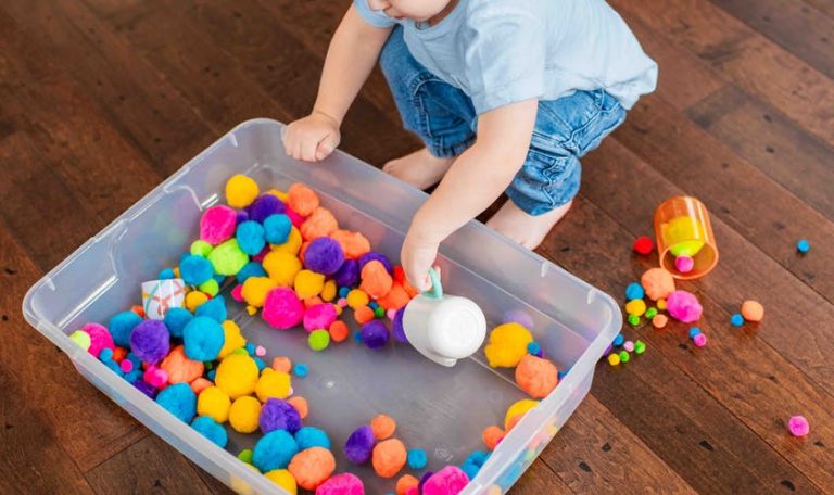 Young child using a cup to scoop up colorful pom poms