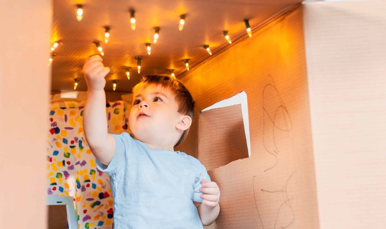 Toddler sitting in a cardboard box that has string lights at the top