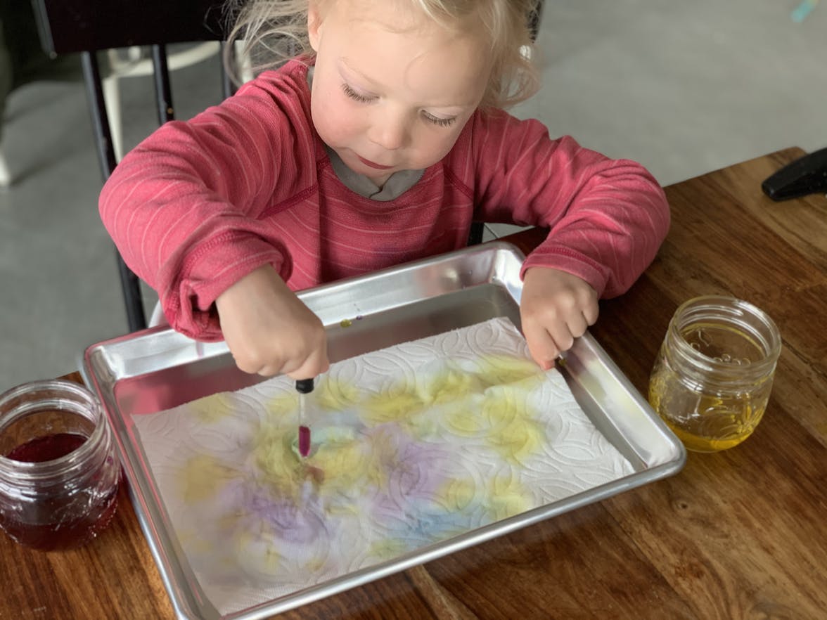 Toddler sitting with a baking sheet and a paper towel dropping colorful water on it
