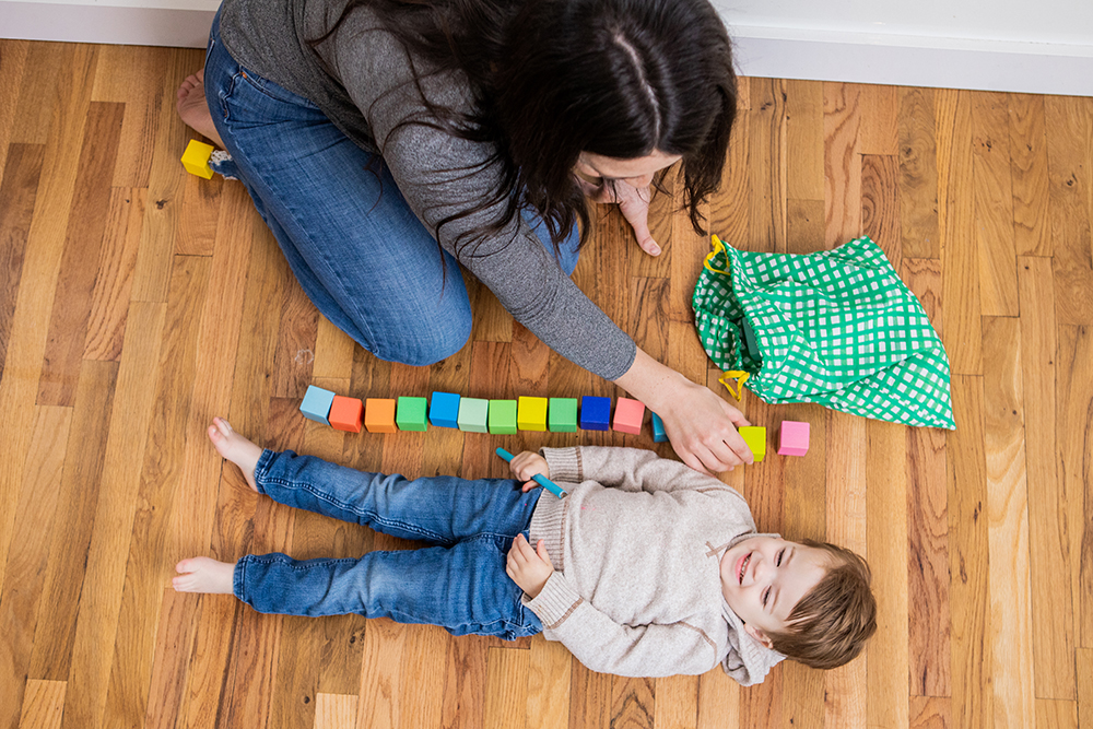 Toddler laying down on the ground next to a stack of blocks laid out by a woman