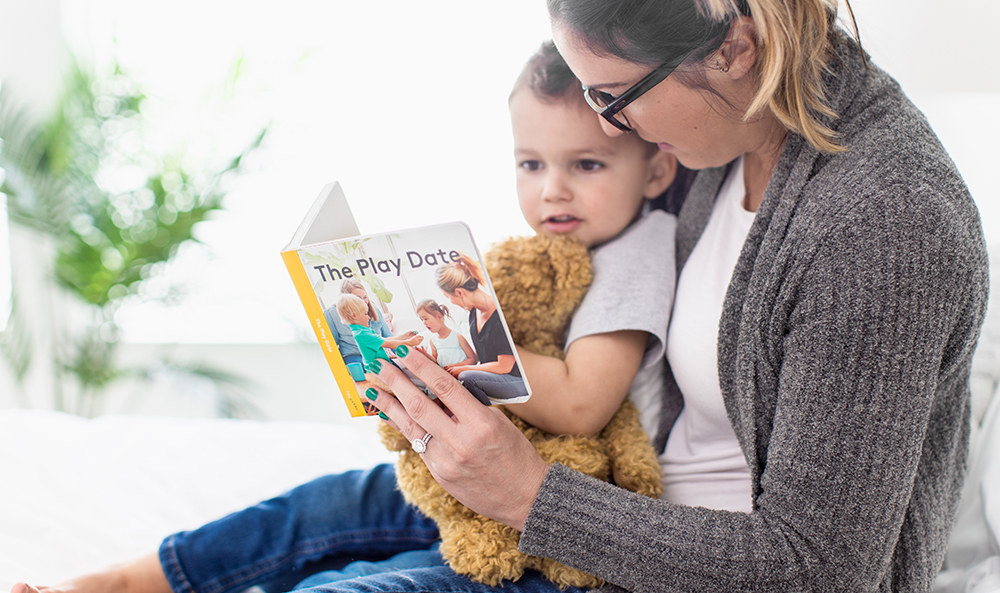 Woman holding toddler while looking at The Play Date book by Lovevery
