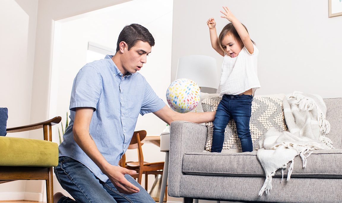 Young child throwing a ball off the couch while being supported by a man