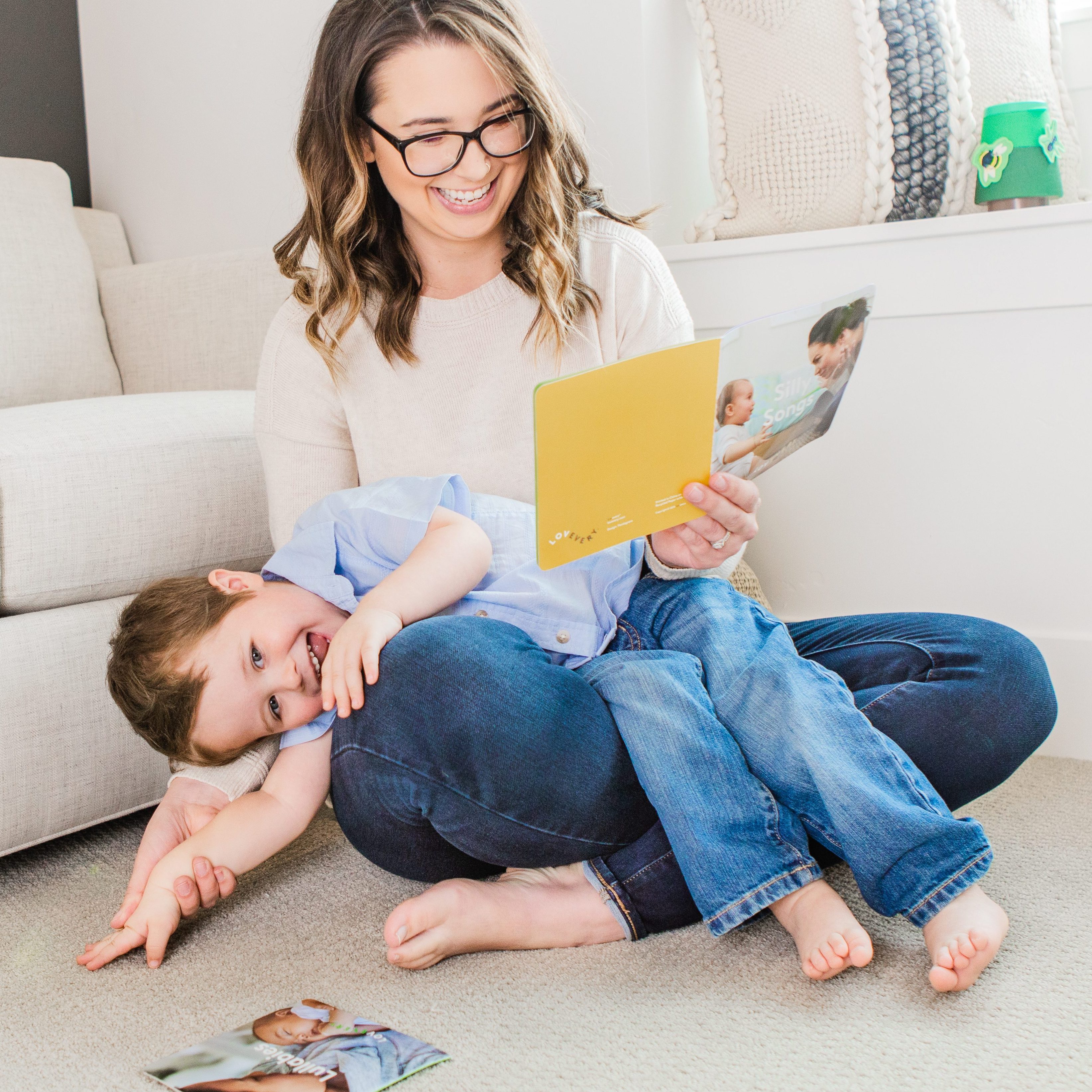 Woman sitting with a toddler on her lap laughing while looking at a book by Lovevery